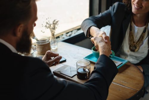 A bearded man shakes hands with a woman in an informal setting whilst enjoying a coffee from a glass beaker